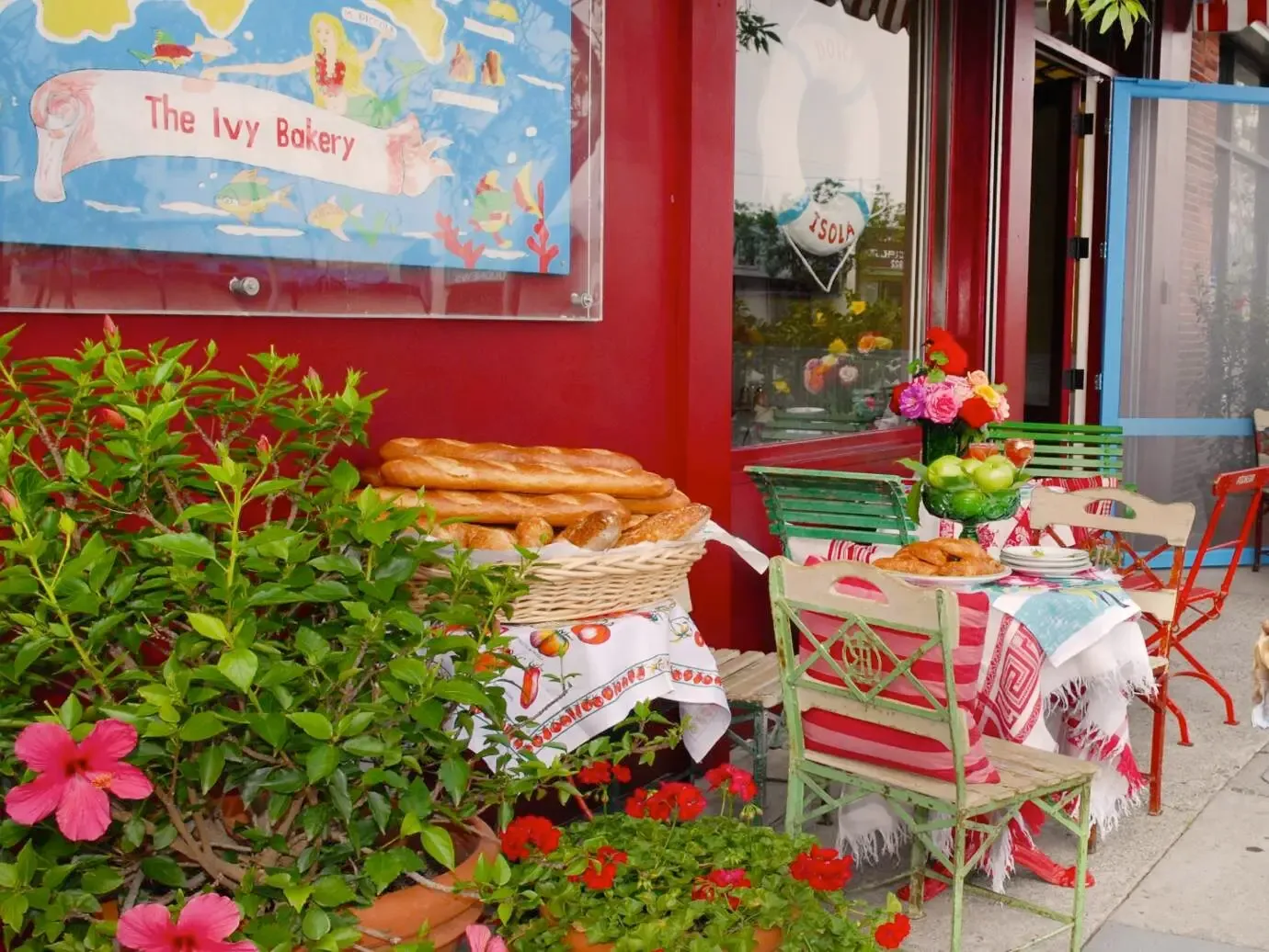 Dolce Isola’s red storefront with baskets of fresh baguettes, café tables, and pots of hibiscus and geraniums on the sidewalk