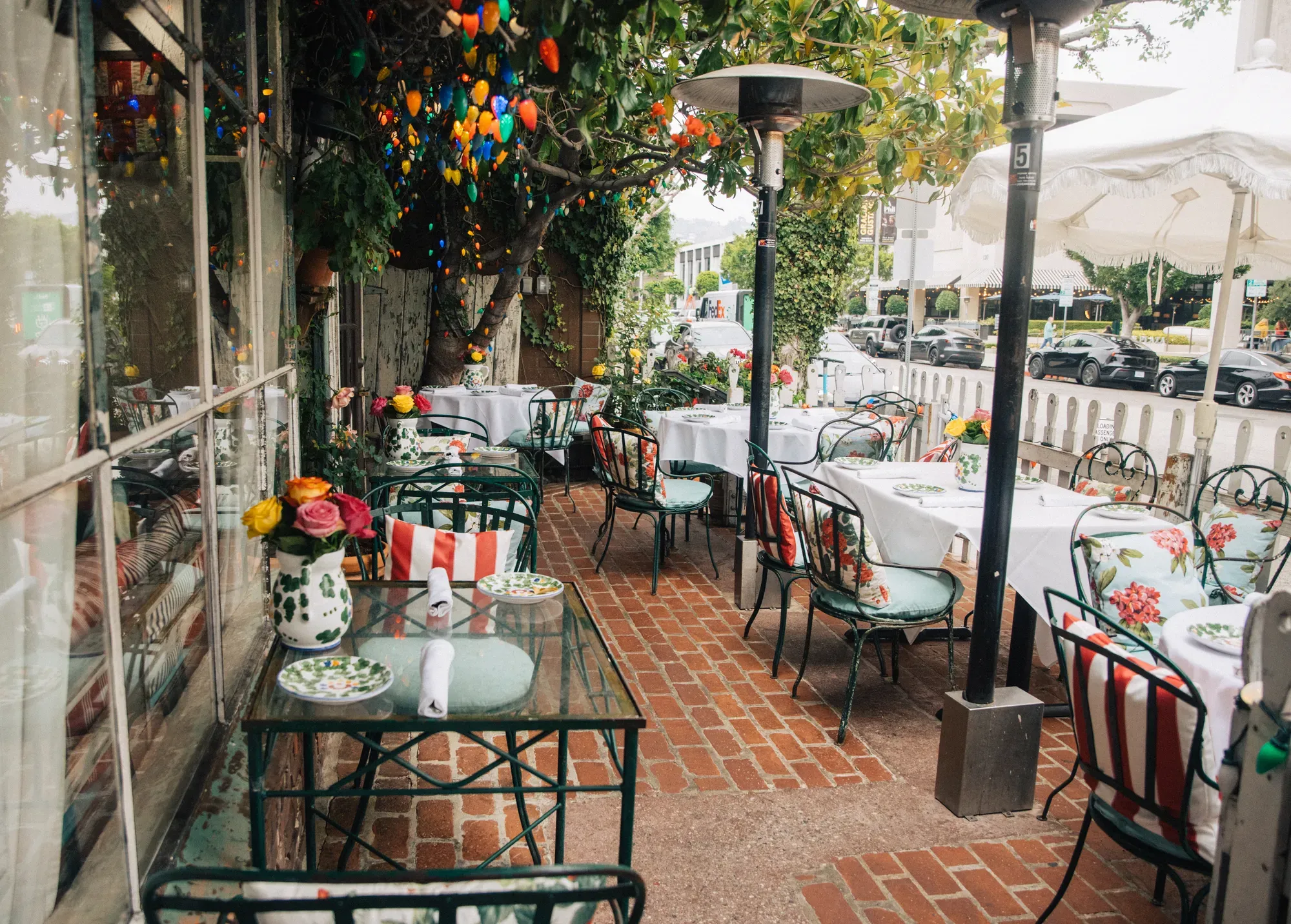 The Ivy’s Side Patio — glass-topped tables set with hand-painted plates and roses, striped and floral cushions on green wrought-iron chairs, string lights in the tree above, and the white picket fence along Robertson Blvd