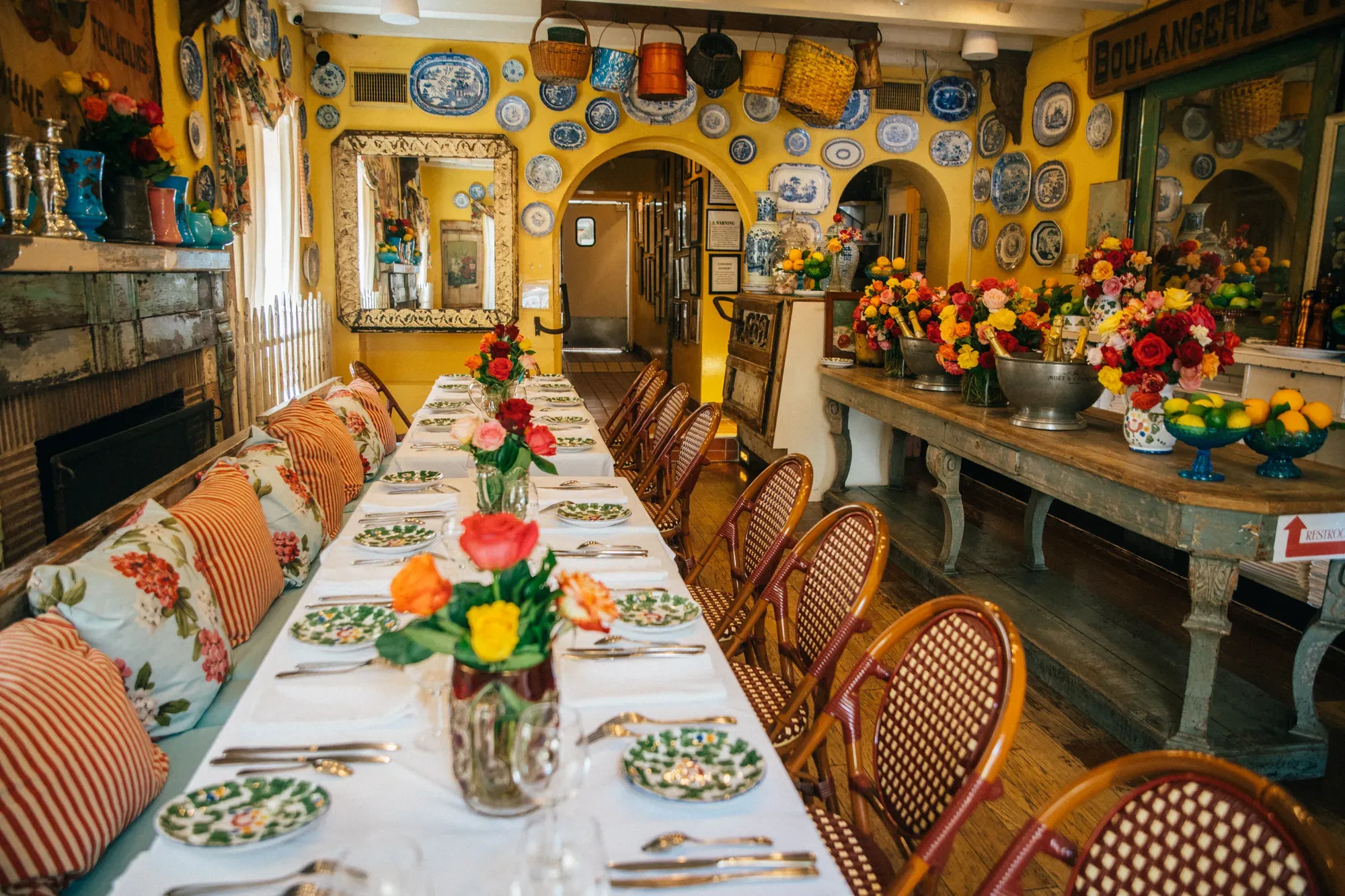 The Lounge at The Ivy set for a private dinner — one long table dressed in white with roses down the middle, rattan café chairs, yellow walls hung with blue-and-white china, baskets overhead, and Champagne on ice